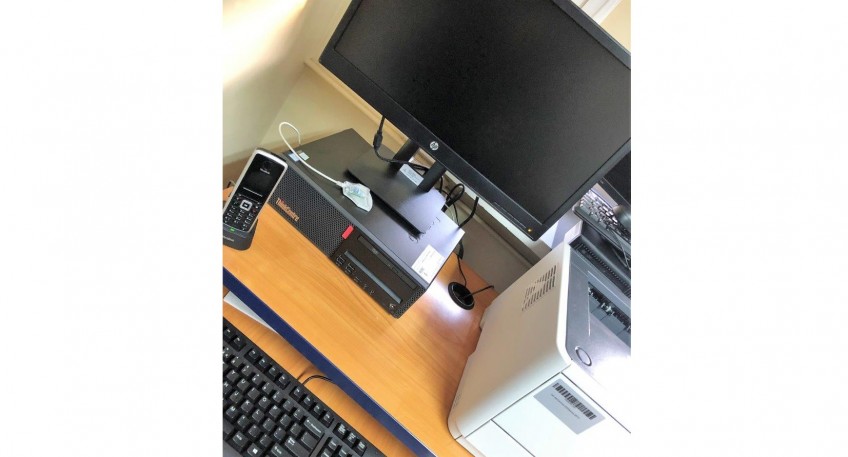 czarno-szary telefon stacjonarny, czarny komputer, czarny monitor, część czarnej klawiatury oraz biało-szara drukarka. Sprzęt stoi na drewnianym biurku.