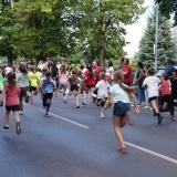 Dzieci w kolorowych strojach, odwrócone tyłem, ścigają się w biegu dziecięcym Szamotulskiej Nocnej Piątki, biegną po ulicy, 