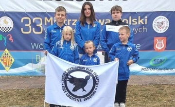 Zawodnicy LUKS „Gry Logiczne” z Wronek na podium na tle banerów imprezy, przed sobą trzymają logo klubu 